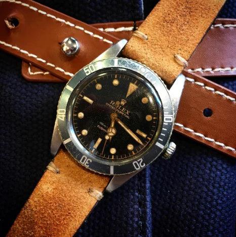 Montre vintage avec cadran noir, boîtier acier et bracelet cuir marron sur tissu bleu.