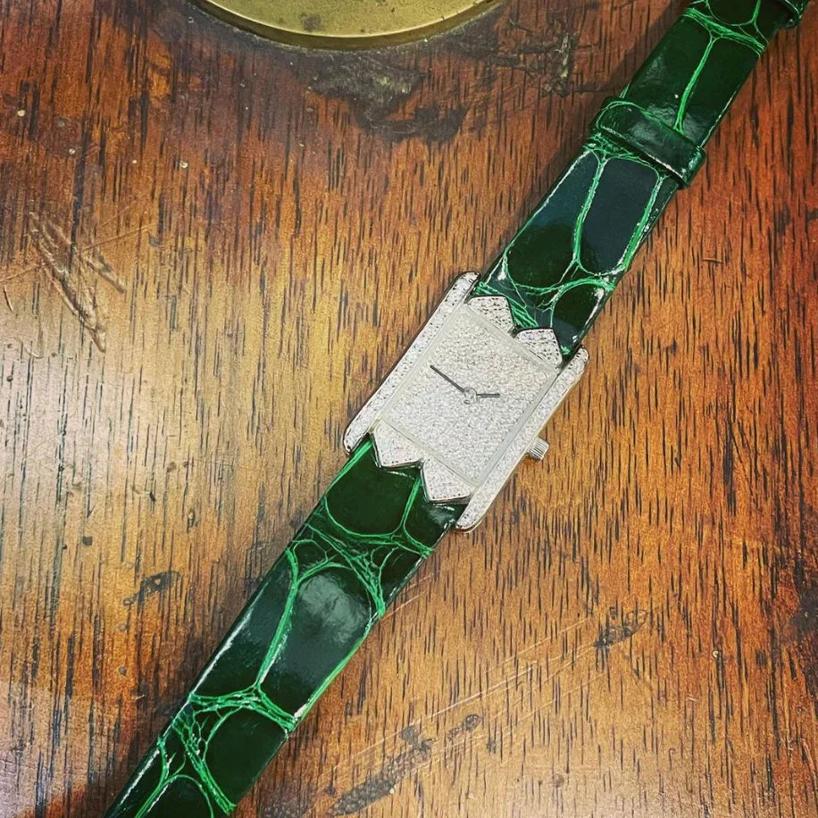 Montre rectangulaire avec cadran diamanté et bracelet en cuir vert sur table en bois.