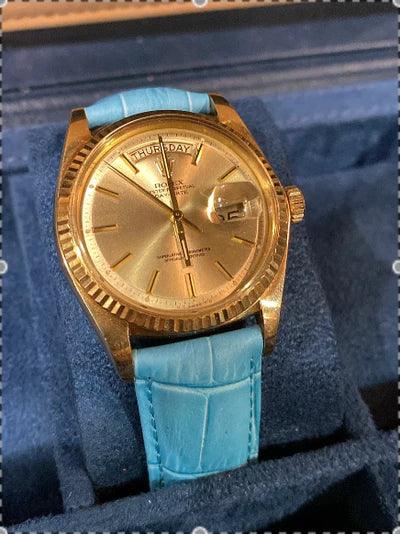 Montre de luxe dorée avec bracelet cuir bleu, cadran jour et date, exposée dans un écrin.