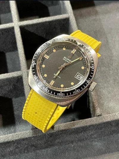 Montre vintage cadran noir avec bracelet jaune, affichage date, posée dans un écrin.